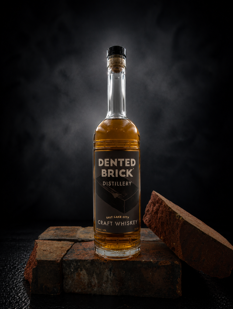 Bottle of Dented Brick Distillery Salt Lake City Craft Whiskey on a brick platform with a dark, smoky background.