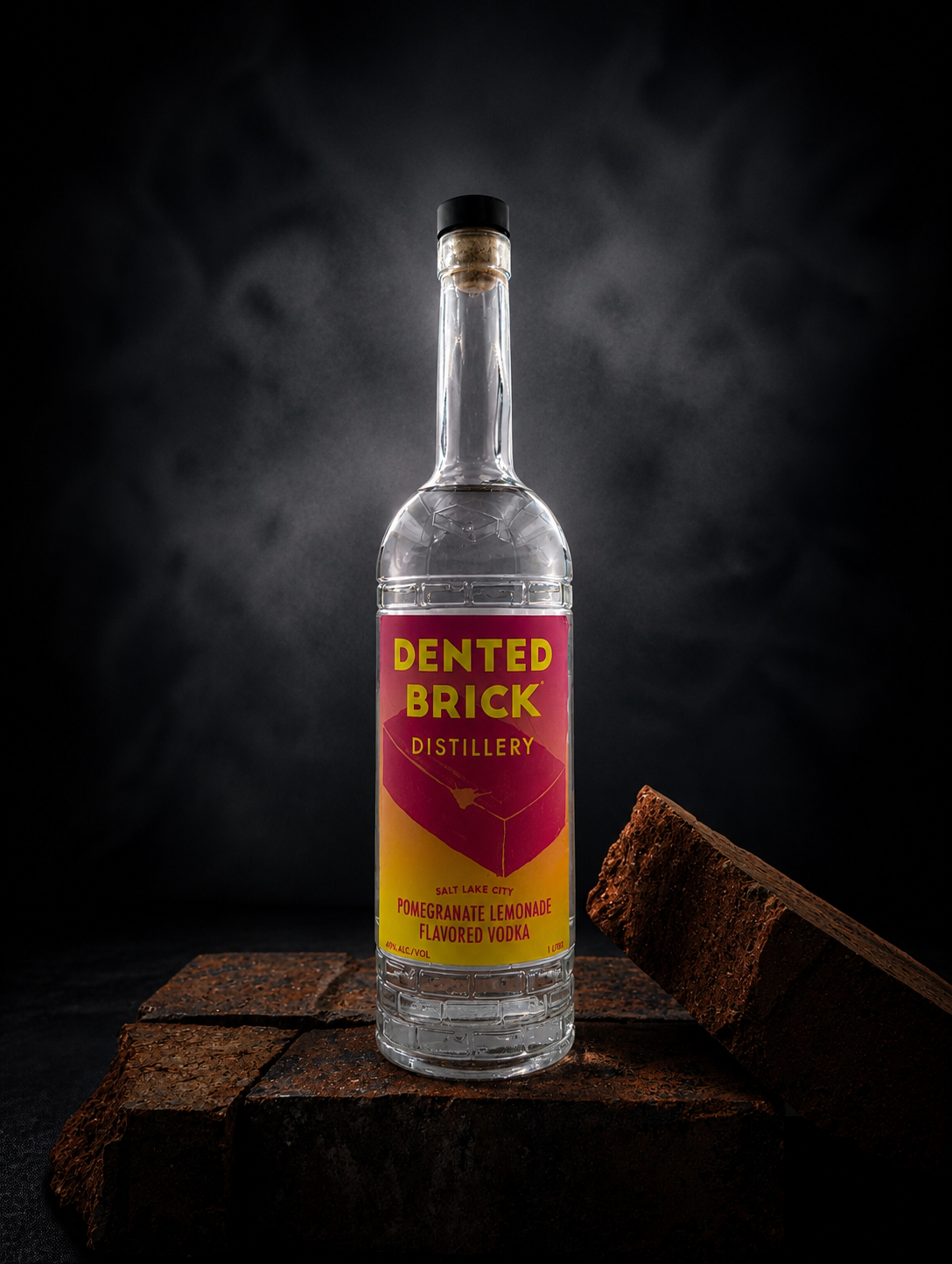 Clear glass vodka bottle with black cap standing on rust-colored bricks in a dark studio, label reads 'Dented Brick Distillery' and 'Pomegranate Lemonade Flavored Vodka' (Salt Lake City).