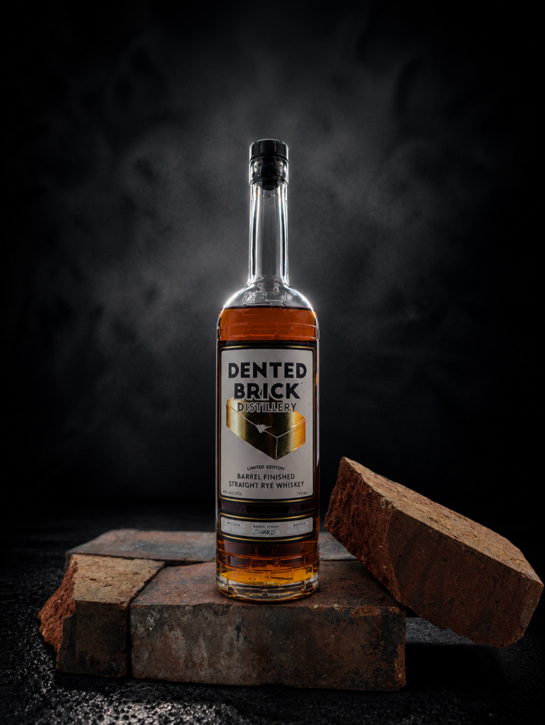 Bottle of Dented Brick Distillery barrel-finished straight rye whiskey on a brick platform with a dark, smoky background.