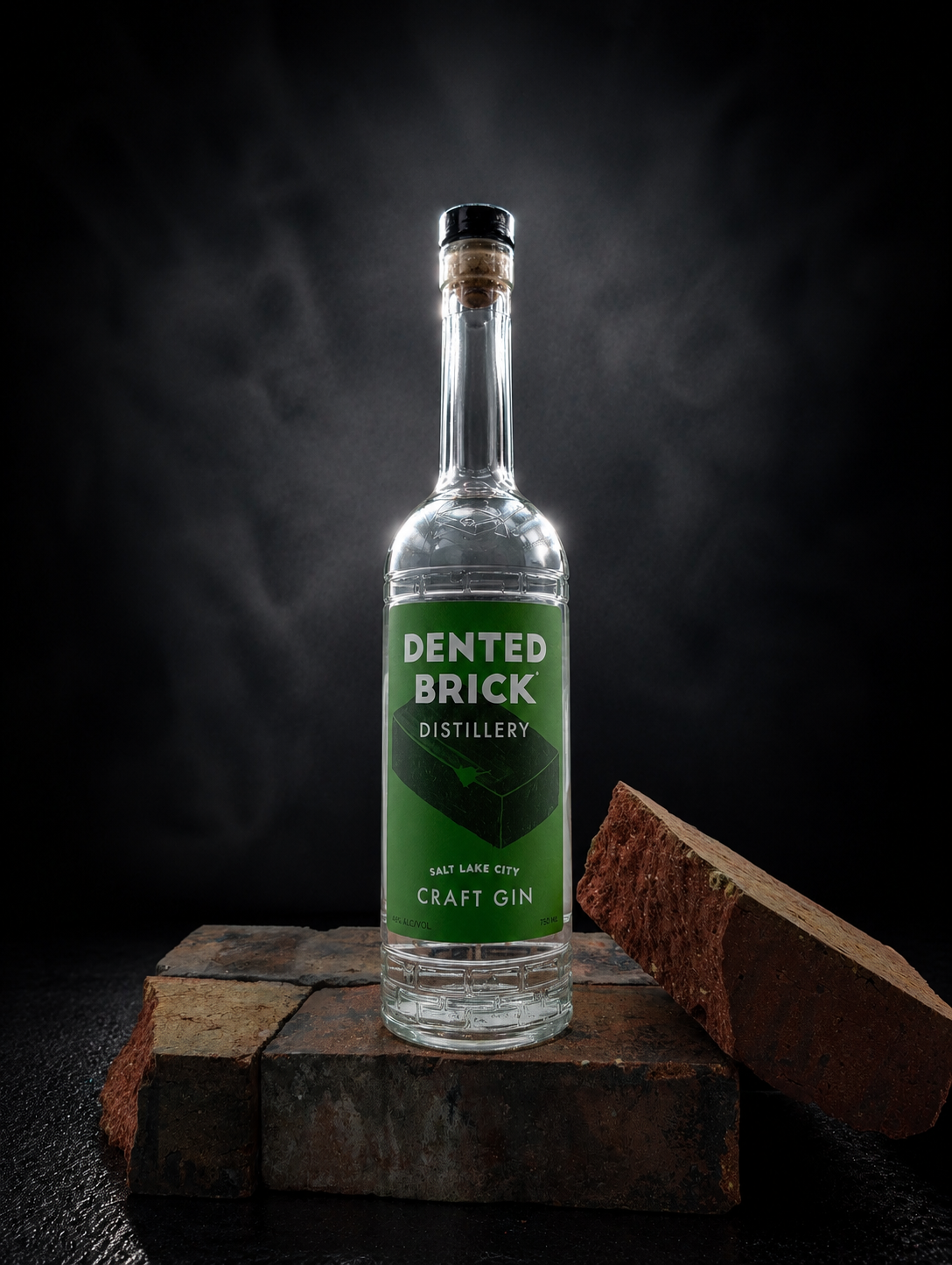 Clear gin bottle from Dented Brick Distillery with a green label on rustic bricks, dark studio background, cork stopper visible.