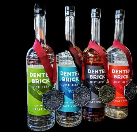 Four Dented Brick Distillery liquor bottles with green, blue, dark red, and orange labels, each tied with a red ribbon and hanging medallion, lined up on a black background.