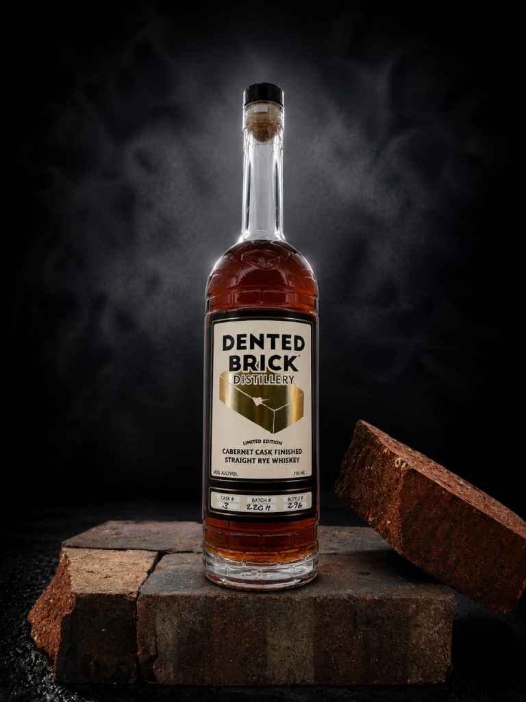 Bottle of Dented Brick Distillery whiskey on a brick pedestal against a dark backdrop, with a broken brick nearby.